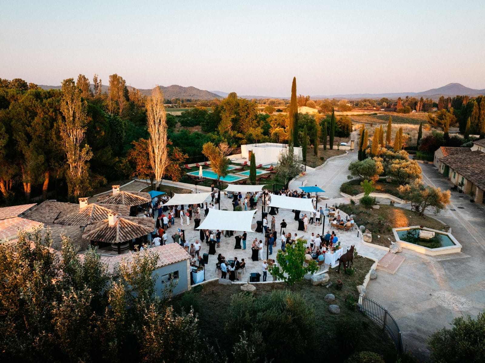 Vue aérienne du Domaine de la Myrtille au coucher du soleil : cocktail sous voiles d'ombrage, piscine et campagne du Luberon